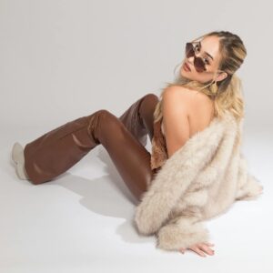 Chic woman wearing sunglasses, furry coat, and brown leather pants on a white background.