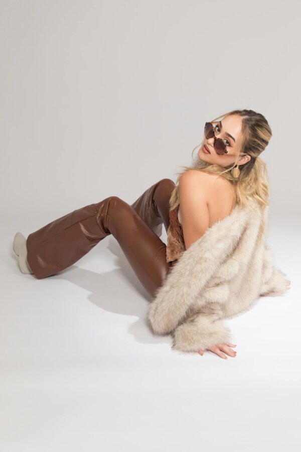 Chic woman wearing sunglasses, furry coat, and brown leather pants on a white background.